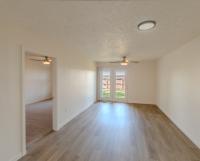Spacious living room with wood-style flooring, neutral walls, and modern light fixtures. French doors open to a fenced outdoor area, and an adjoining doorway leads into the bedroom.